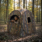 A camouflage pop-up hunting blind set in a wooded forest, with a person in camo leaning out of the opening holding a rifle.