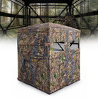 A camouflage hunting blind with a realistic woodland print, shown in front of a forest backdrop with a hunter visible inside. The product features a multi-view design with window cutouts, set against a white foreground gradient.