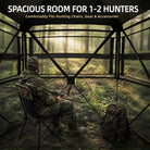 A camouflage hunting chair with a camouflage tree-bump stand inside a screened forest hunting blind, with a man in camouflage sitting in the chair and looking out toward a sunlit woods background. Text at the top reads: 'SPACIOUS ROOM FOR 1-2 HUNTERS' and 'Comfortably Fits Hunting Chairs, Gear & Accessories'.