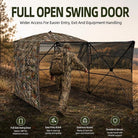 A camouflage hunting blind with a large open swing door is shown set in an outdoor field. A person in camouflage walks through the open doorway, demonstrating easy access.