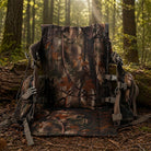 A camouflage backpack laid open on the forest floor, with a matching camouflage floor pad or mat placed in front. The setting is a wooded outdoor scene with trees, moss, and sunlight filtering through the woods.